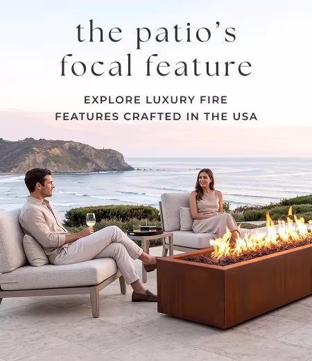Four people gather and converse on a luxury outdoor patio around a long, rectangular weathering steel fire pit with a coastal ocean backdrop at sunset. Image Text: the patio's focal feature EXPLORE LUXURY FIRE FEATURES CRAFTED IN THE USA